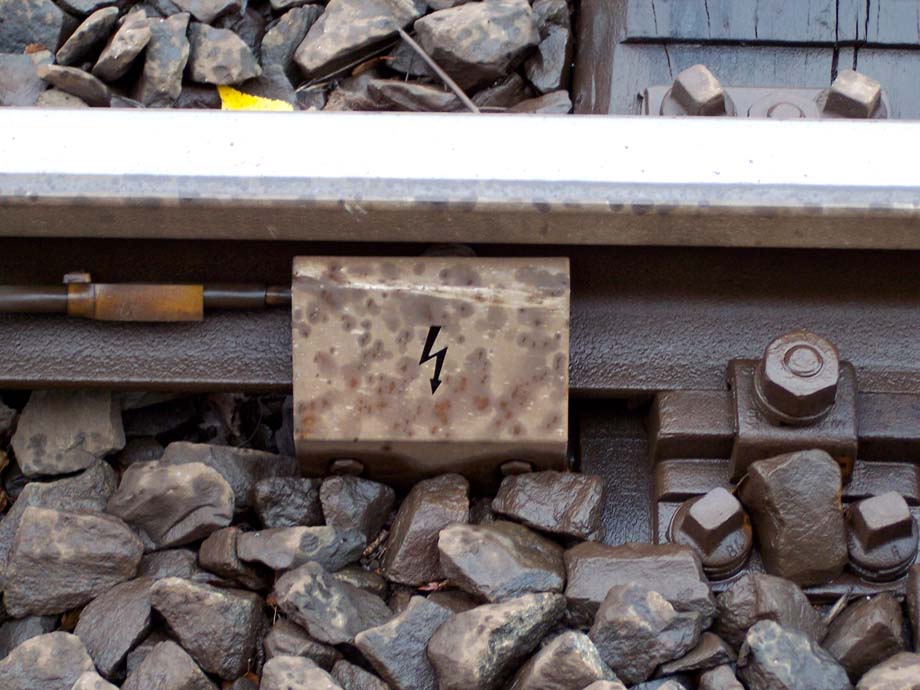 electricity warning symbol (lightning bolt) where cable connects to rail on ballast; wet from rain drops | bolt of Iuppiter fulgurans/pluvialis; photo